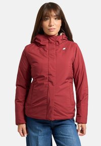 Giacca imbottita rossa con colletto alto e cappuccio, dotata di un piccolo logo sul petto e tasche laterali. Texture sintetica liscia.