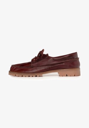 Jambes portant une jupe en velours côtelé marron, des chaussettes bordeaux avec un bord rose et des chaussures en cuir marron à lacets, debout sur un sol beige.