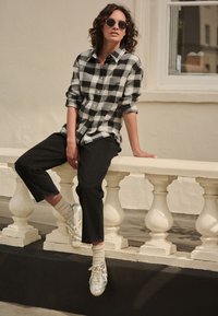 Černobílá károvaná flanelová košile, černé džíny, stříbrné tenisky, světlé ponožky; sedí na ozdobném zábradlí.