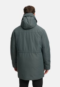 Vadderad kappa i mörkgrönt med huva, som har en linjeseam vid midjan och en slät textur. Baksidan visar en enkel och funktionell design.