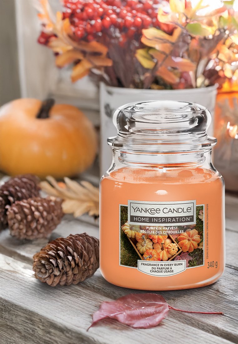Świeca Yankee Candle w przezroczystym słoju ma zapach zbiorów dyni. Świeca jest pomarańczowa, ozdobiona etykietą i stoi w otoczeniu szyszek oraz jesiennych liści.