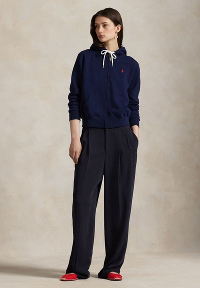 Γυναίκα που φοράει ναυτικό μπλε φούτερ με φερμουάρ, φαρδιά μαύρη παντελόνια και κόκκινα παπούτσια χωρίς κορδόνια, στέκεται με τα χέρια στις τσέπες μπροστά σε ουδέτερο φόντο.