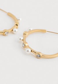 Guld hoop øreringe prydet med små hvide perler og klare ædelstene på en hvid baggrund.