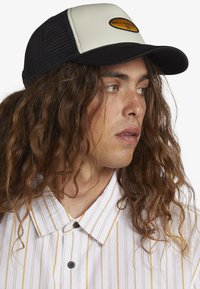 Black and cream trucker cap with mesh sides, featuring a circular logo patch. Paired with a white shirt that has yellow vertical stripes.