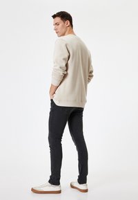 Sudadera beige con puños y dobladillo acanalados, combinada con jeans negros y zapatillas blancas con suela marrón. El modelo está ligeramente girado.
