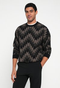 Brązowy i czarny sweter we wzór jodełki, z prążkowanymi mankietami i kołnierzem, noszony z czarnymi spodniami.