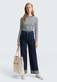 Pasiasta koszula z długim rękawem, ciemnoniebieskie dżinsy o wysokim stanie z szerokimi nogawkami i podwiniętymi mankietami, torba na ramię z naturalnej tkaniny oraz białe trampki.