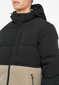Black and beige padded jacket with a zippered hood, featuring a vertical chest pocket and side zipper pockets. Smooth texture and geometric stitching.