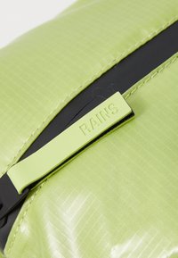 Saco impermeável verde com acabamento texturizado, apresentando um detalhe preto e uma etiqueta com o logotipo "RAINS" visível. Material liso e brilhante.