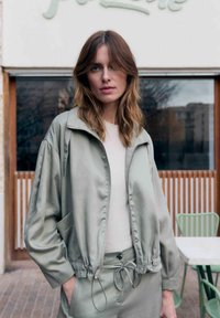 Vrouw met schouderlang bruin haar die een lichtgrijze zip-up jas en bijpassende broek draagt, staand buiten nabij een tafel en stoelen.