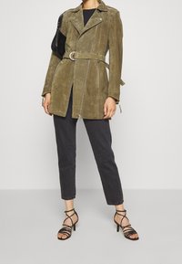 Veste en daim verte olive ceinturée avec un design de moto, détails de fermeture éclair argentés et manches longues, assortie à un jean noir et des talons à lanières.