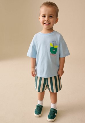 Smiling toddler wearing a light blue T-shirt with a green apple graphic, striped green and white shorts, white socks, and green sneakers.