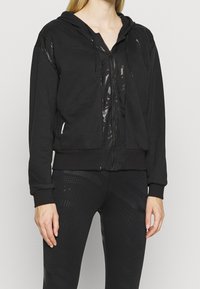 Un sweat à capuche zippé noir avec un panneau avant brillant et texturé, un capuchon à cordon de serrage et des poignets côtelés, associé à un legging noir à motifs assortis.