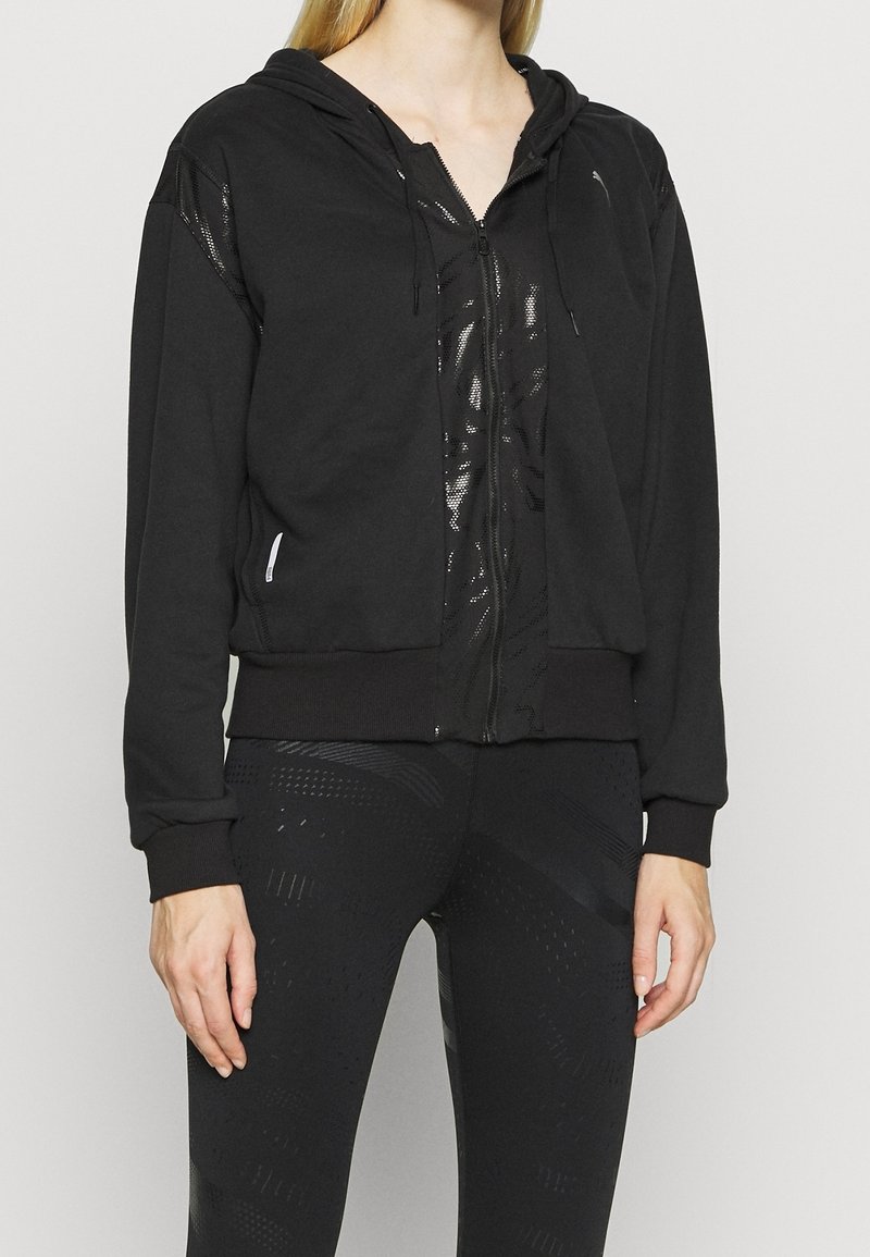 Un sweat à capuche zippé noir avec un panneau avant brillant et texturé, un capuchon à cordon de serrage et des poignets côtelés, associé à un legging noir à motifs assortis.