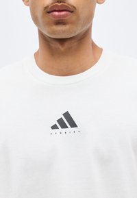 Gros plan sur un homme portant un t-shirt blanc à col rond avec un logo Adidas noir et le mot « RUNNING » dessous.