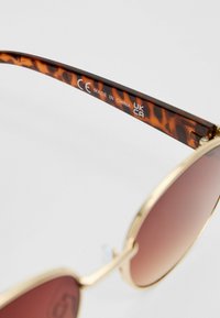 Lunettes de soleil rondes avec une monture en métal doré et des verres teintés en ambre. Les branches présentent un motif écaille de tortue avec des détails gravés.