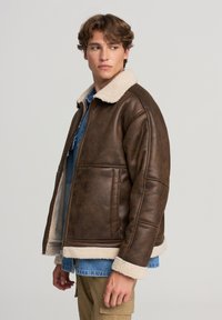 Brązowa kurtka z faux skóry z kremowym kołnierzem z owczej wełny, zamkiem z przodu i bocznymi kieszeniami; ma fakturowane wykończenie i luźny krój.