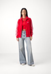 Giacca da track in poliestere rossa con strisce bianche, indossata sopra un crop top rosso, abbinata a jeans larghi azzurri e sneaker bianche.