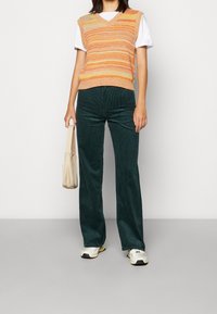 Mångfärgad stickad väst med v-ringning, tillsammans med gröna kordfloddsbyxor med vida ben. En ljusbeige handväska och ljusa sneakers fullföljer looken.