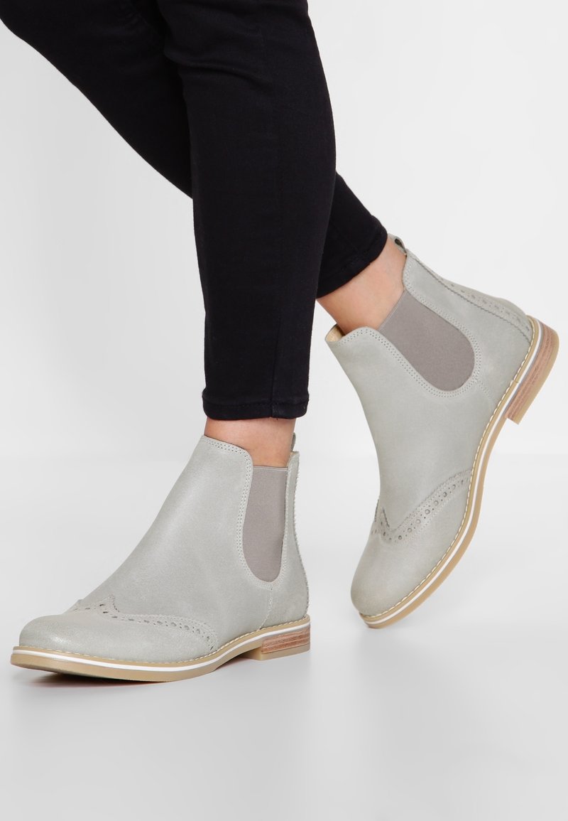 Personne portant des bottines en cuir gris clair avec des empiècements élastiques latéraux et des semelles en bois, associées à un pantalon noir ajusté.
