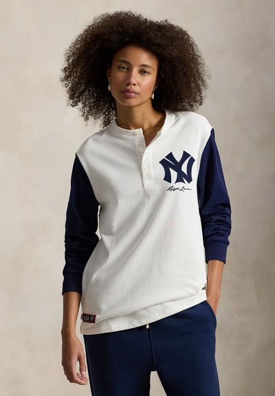 Chemise henley à manches longues blanche et marine avec texture côtelée, présentant un logo et une signature marine sur la poitrine, ainsi qu'une accent rouge à l'ourlet.