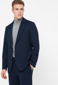 Jovem vestido com um fato azul marinho e um suéter de gola alta cinza, de pé com uma mão no bolso, olhando para o lado.