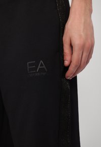Gros plan d'une main reposant à côté d'un pantalon noir avec un logo EA Emporio Armani discret et une bande latérale texturée.