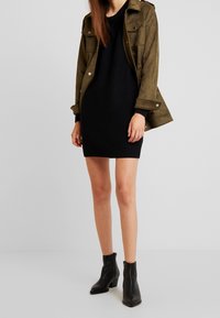 Giacca in suede verde oliva sopra un vestito nero a maglia, che mostra una vestibilità rilassata e un tessuto testurizzato. Stivaletti neri alla caviglia con tacco basso.