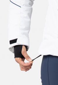 Bílá voděodolná bunda s černými elastickými manžetami. Ruka upravuje šňůrku na spodním lemu. Hladká textura, přiléhavý střih, minimální akcenty.