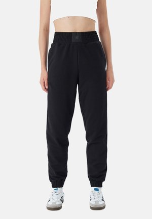 Schwarze Jogginghosen mit einem rüschierten Bund, schmal zulaufenden Bündchen und seitlichen Taschen, kombiniert mit weißen Sportschuhen und gemusterten Socken.