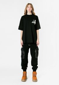 Camiseta negra de gran tamaño con gráfico lateral, combinada con pantalones cargo negros con bolsillos con cremallera y botas marrones con suela gruesa.