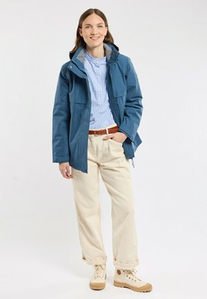 Veste imperméable bleue avec doublure de capuche grise, chemise à rayures à boutons, pantalons beiges et bottes beige clair avec semelles texturées.