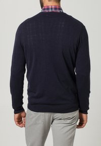 Marineblauer Pullover mit strukturiertem Finish, Rundhalsausschnitt und gerippten Bündchen. Getragen über einem karierten Hemd mit roten Akzenten.