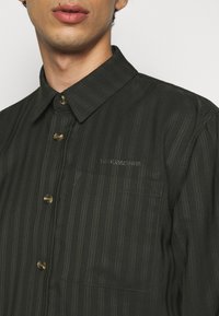 Mörkgrönrandig skjorta med knappar och klassisk krage, featuring bruna knappar, ficka på bröstet och broderat varumärke.