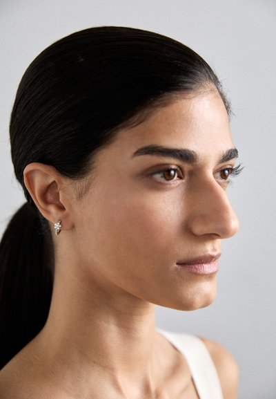 Gold earring with a floral design featuring small clear crystals, worn on the left ear. Smooth, dark hair pulled back. Neutral background.