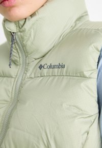 Zeleni puf prsluk marke Columbia, s visokim ovratnikom, zatvaranjem na patentni zatvarač i suptilnim kariranim uzorkom na glatkoj, quilted površini.