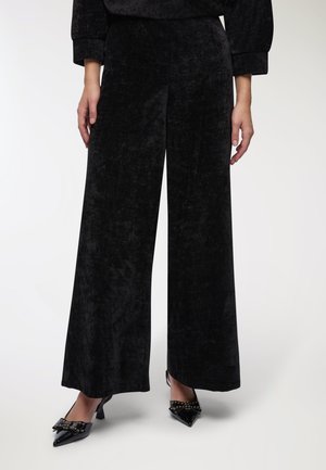 Pantaloni in velluto neri a gamba larga con una texture morbida, abbinati a décolleté nere a punta con embellishments chiodati.