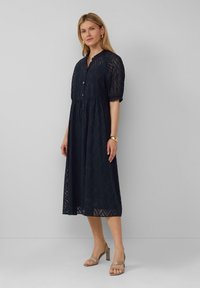 Robe midi bleu marine avec un motif en vagues texturé, manches courtes, fermeture à boutons et taille froncée. Accompagnée de sandales à talons de couleur claire.
