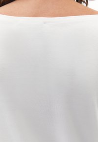 Hvid bomuldsbluse med en bred udskæring, glat tekstur og minimal syning detalje bagpå, der fremhæver et enkelt, grundlæggende design.