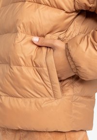 Doudoune légère couleur beige, avec un tissu matelassé lisse, une poche latérale et des poignets élastiques pour un ajustement confortable.