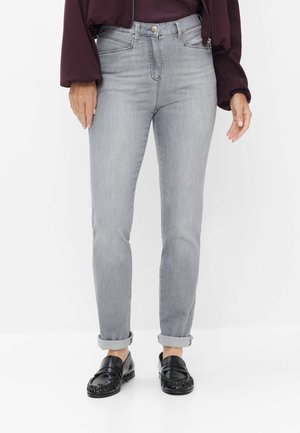 Person iført lysegrå bukser med opsmøg, sorte loafers og en mørk langærmet top, stående mod en hvid baggrund.