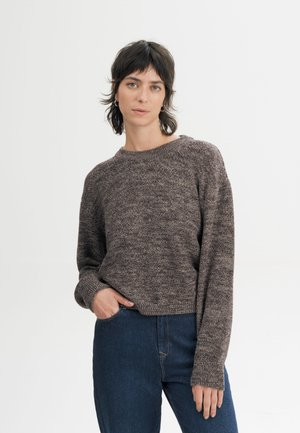 Persoon met kort donker haar, gekleed in een bruine gebreide trui en blauwe spijkerbroek met één hand in de zak, staand tegen een lichte achtergrond.