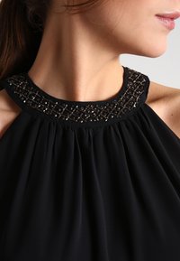 Sort halterneck-top med plisseret stof og detaljer med perler ved udskæringen, der har en glat tekstur og subtil glans på accenterne.