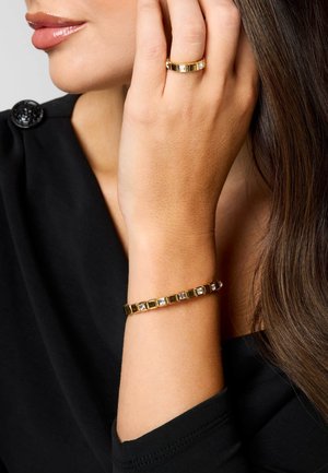 Frau trägt goldenen Ring und passendes Armband mit quadratischen klaren Steinen, Hand ruht nahe am Gesicht, trägt schwarzen Oberteil.