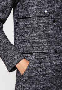 Mano in tasca di un cappotto grigio scuro con texture, bottoni neri e tasca a pattina sul petto, che mostra i dettagli del tessuto intrecciato e della manica.