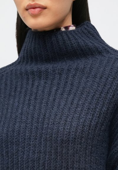 Γυναίκα που φοράει παχιά, �σκούρα μπλε πλεκτή μπλούζα με ριμπ σχέδιο και ζιβάγκο, με μια υποψία από ρούχο με σχέδιο από κάτω.