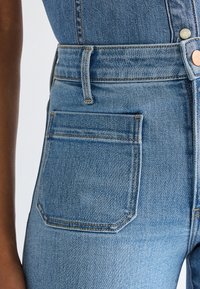 Detail světle modrých džín s viditelným zadním kapsou, poutkem na pásek a měděným knoflíkem v pase vedle džín s tmavou pokožkou ruky.