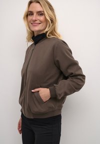 Veste bomber marron zippée avec une finition texturée, des manches côtelées et des poches latérales. Portée par-dessus un col roulé noir, présentant un design décontracté.