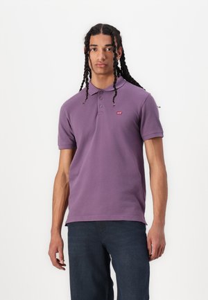 Violetinio medvilninio polo marškinėlių su trimis mygtukais ir mažu raudonu logotipu kairėje krūtinės pusėje derinys su mėlynomis džinsomis.