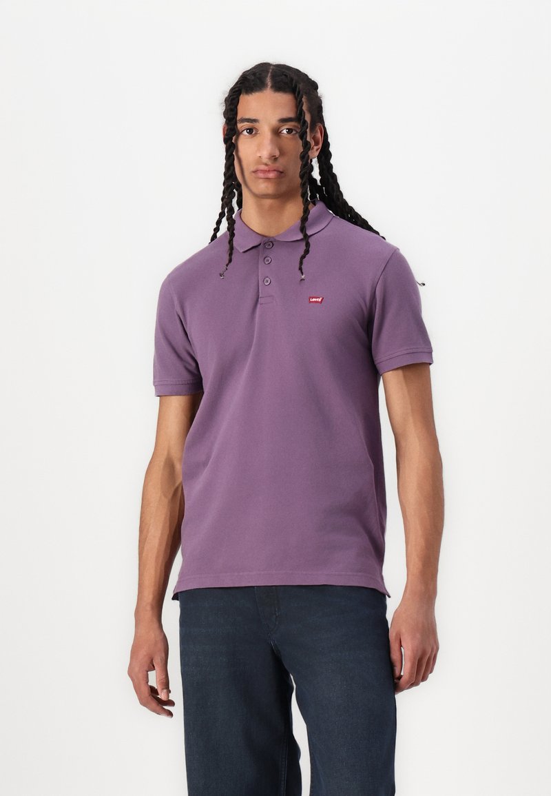 Tricou polo violet din bumbac, cu o deschidere cu trei butoane și un mic logo roșu pe pieptul stâng, asortat cu blugi albaștri.